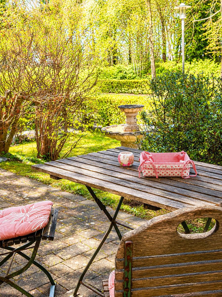 Holztisch mit Kissen und Korb im Garten. Hintergrund: Brunnen und grüne Hecken.