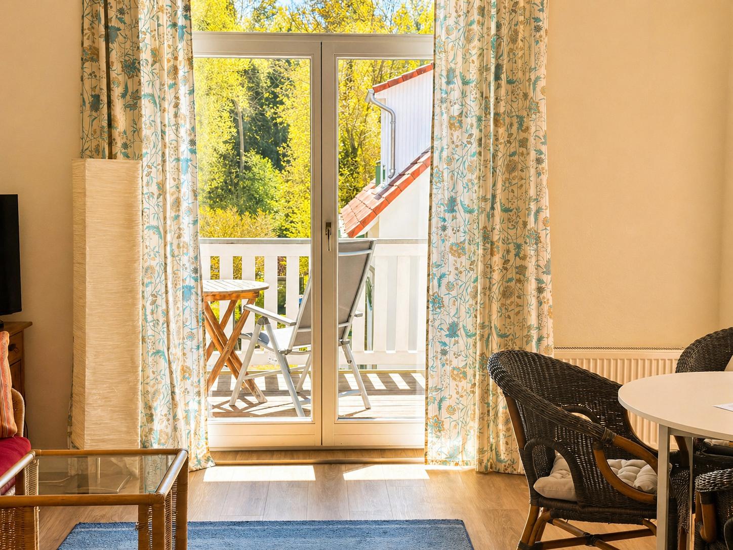 Wohnraum mit Blick auf Balkon, Tisch und Stühle. Vorhänge und Holzboden sichtbar.