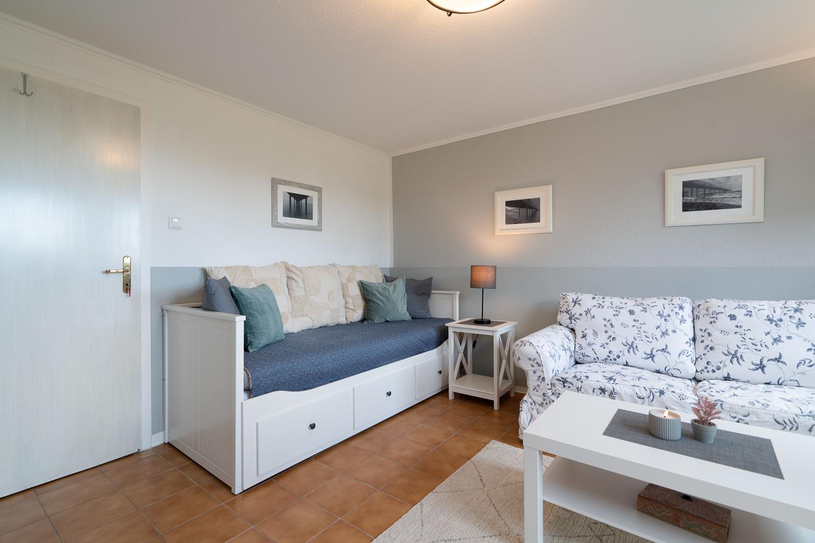 Living room with sofa, bench, and table. Walls painted gray-white. Tile floor and pictures on the wall.