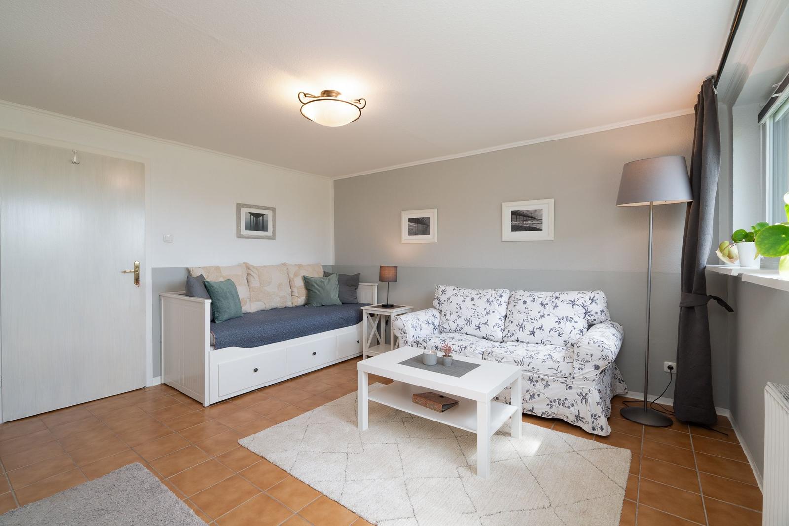 Living room with sofa, bench, table, and floor lamp. Gray-white walls, tiled floor.