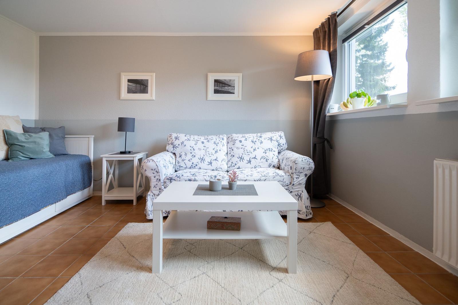 Living room with sofa, coffee table, and bed. Window with curtains and plants.