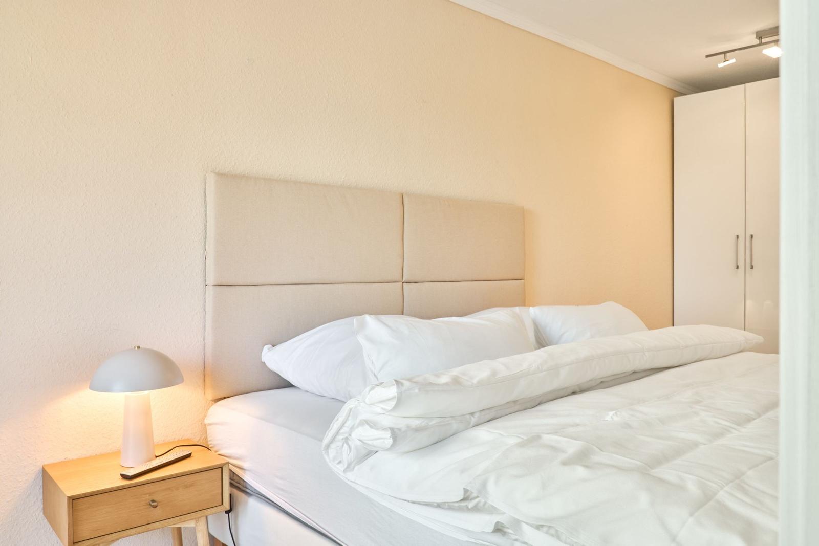 Schlafzimmer mit Bett, Nachttisch und Schrank. Wandbeleuchtung und weiße Bettwäsche.