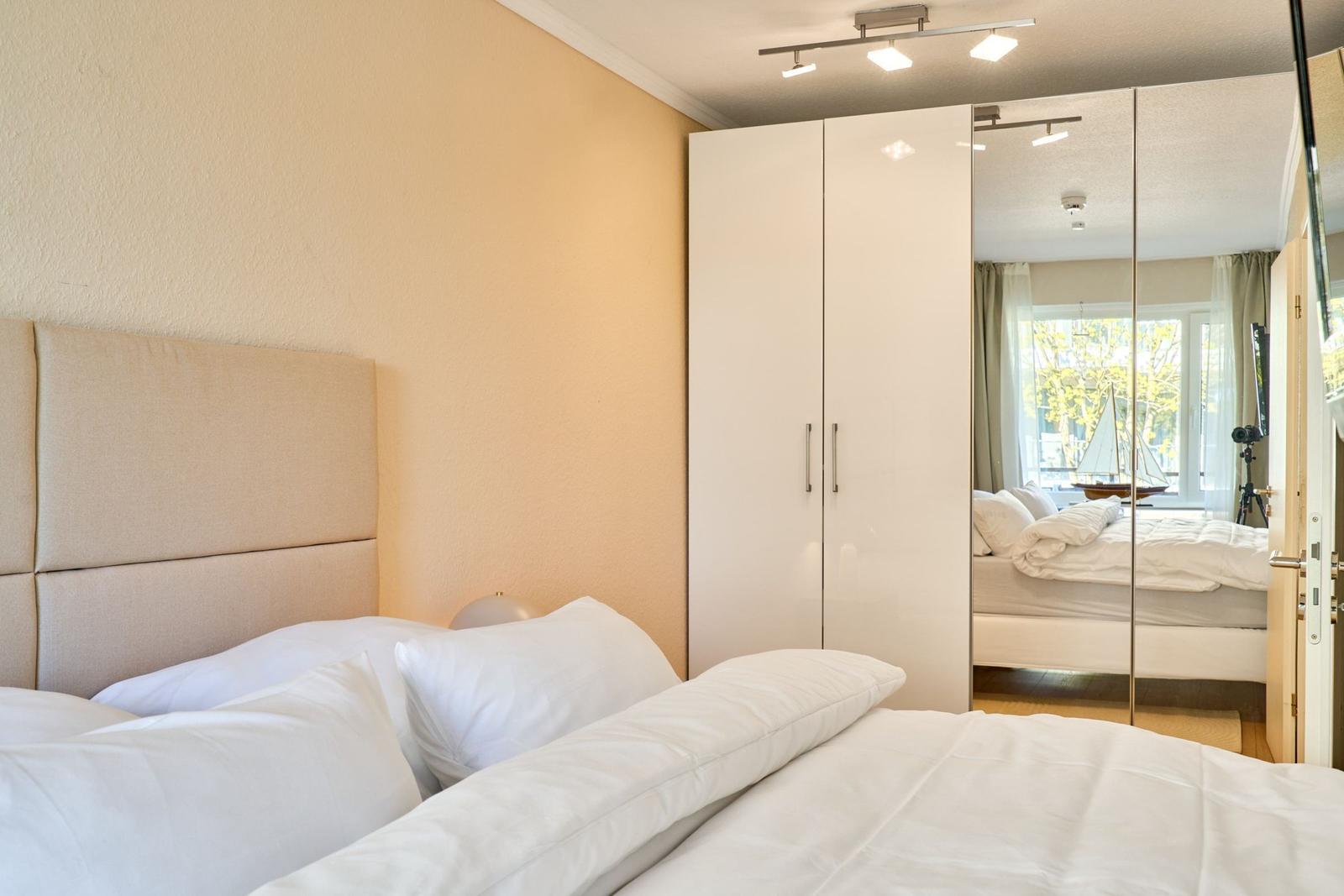 Schlafzimmer mit Bett, weißen Kissen und Spiegelschrank. Durchgang zu Wohnbereich mit Fenster.