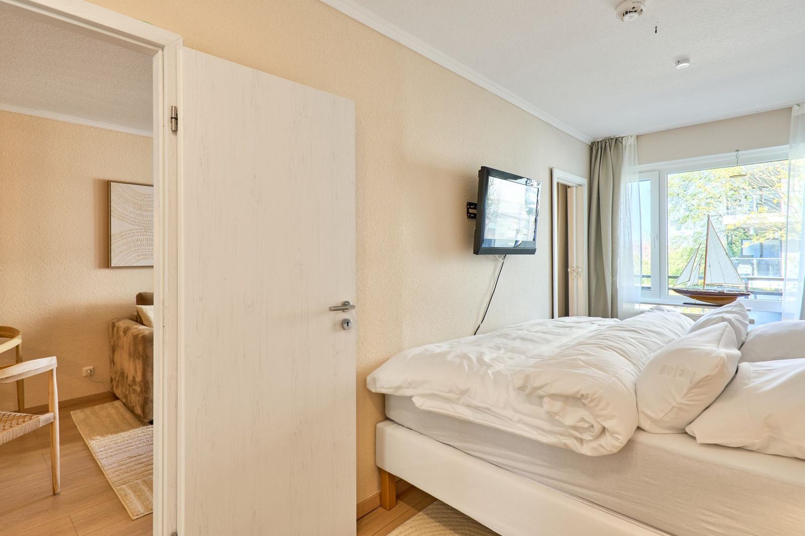 Schlafzimmer mit Bett, Wandfernseher und Blick auf Balkon mit Segelbootmodell.