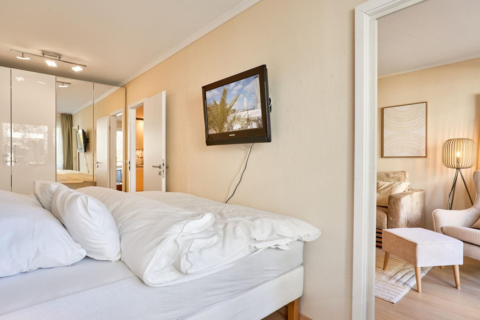 Schlafzimmer mit Bett, TV und Blick in Wohnbereich mit Sitzmöbeln.