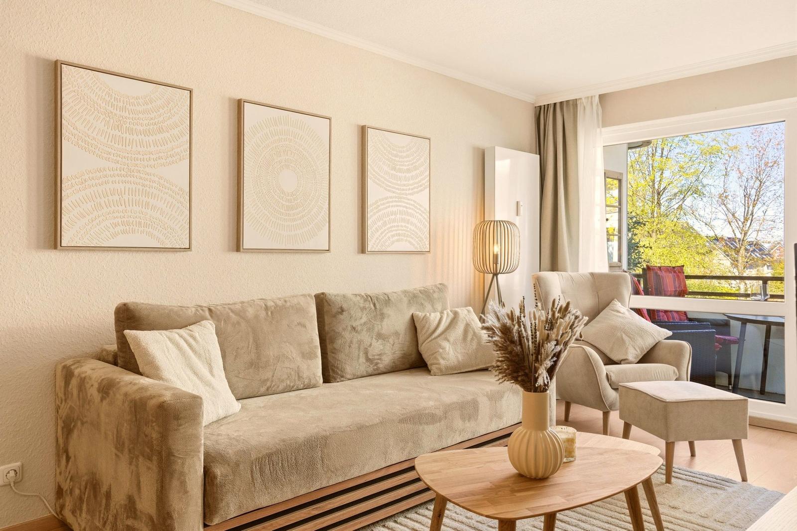 Wohnzimmer mit Sofa, Sessel und Tisch. An der Wand hängen drei Bilder. Durch das Fenster ist eine Terrasse zu sehen.