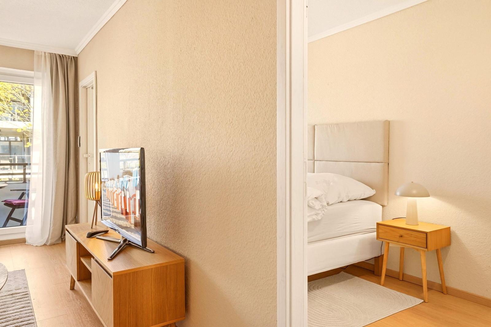 Ein Zimmer mit Bett und Nachttisch. Fernseher auf Holzregal. Fenster mit Vorhängen.