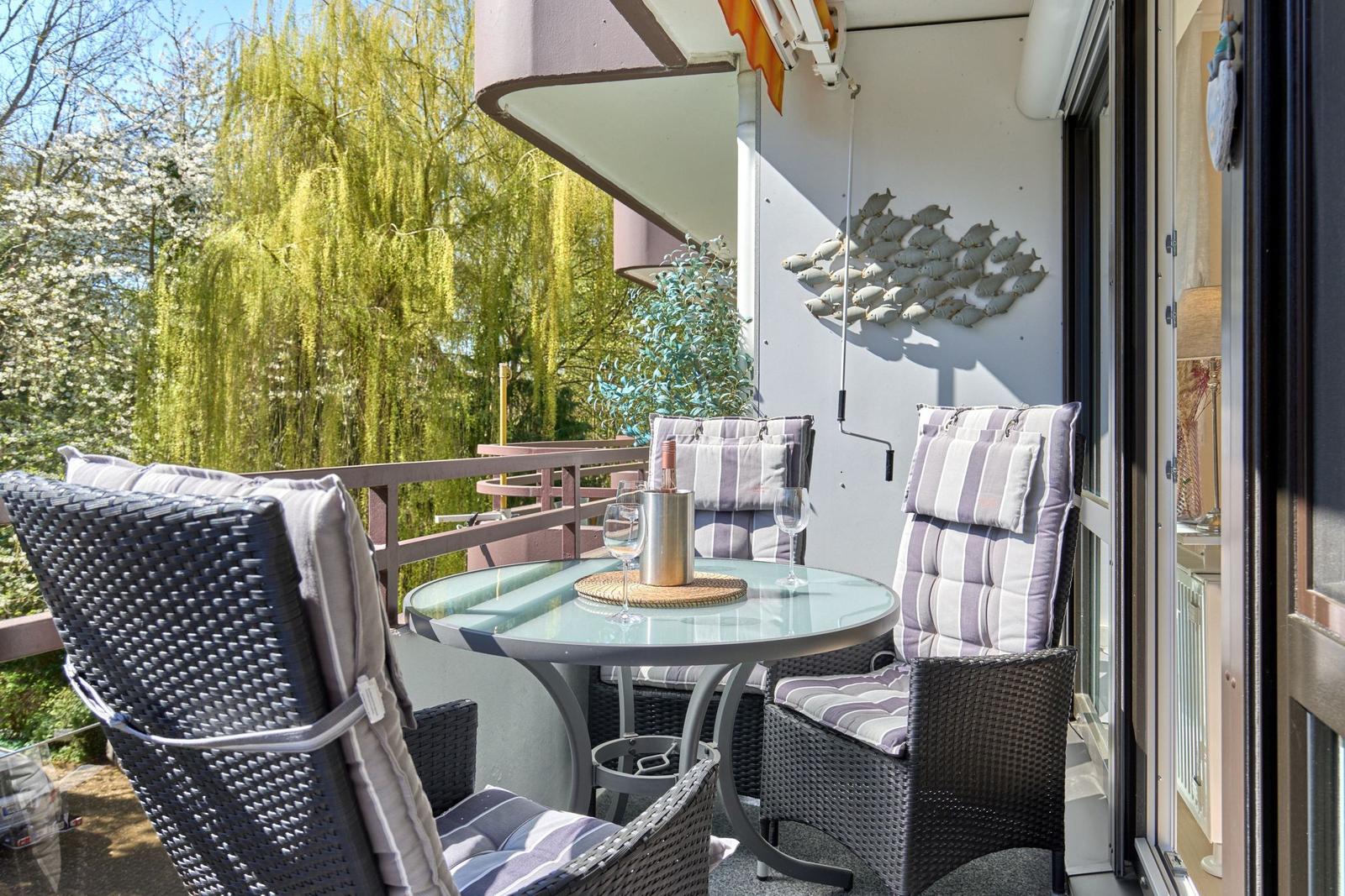 Terrasse mit Sitzgruppe, Tisch und Weinflasche vor grüner Vegetation.