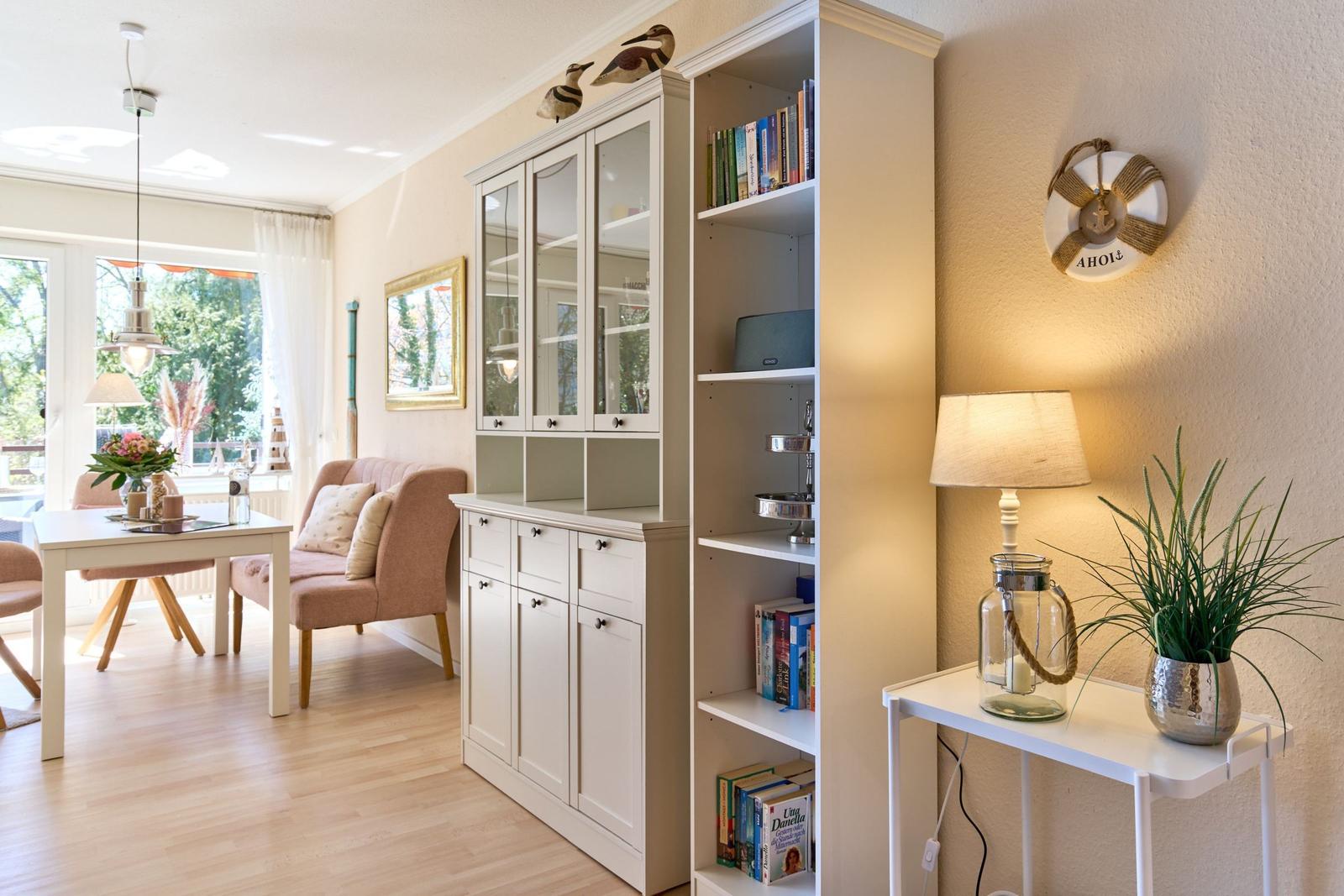 Wohn- und Essbereich mit Bücherregal, Sitzbank und Tisch. Wanduhr und Lampe sind sichtbar.