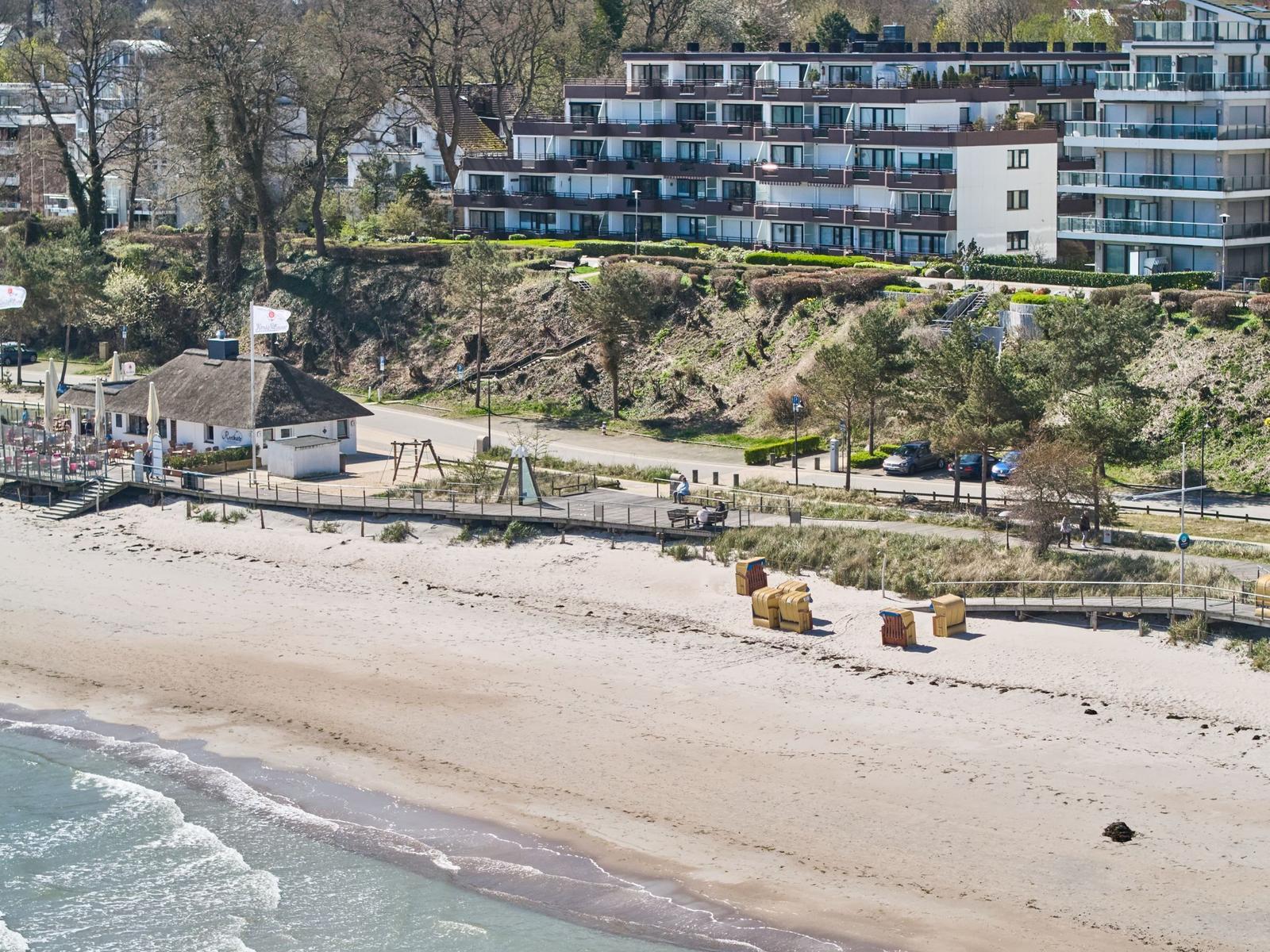 Strand mit Sand, Wellen und Strandkörben. Im Hintergrund Wohnhäuser und Bäume.