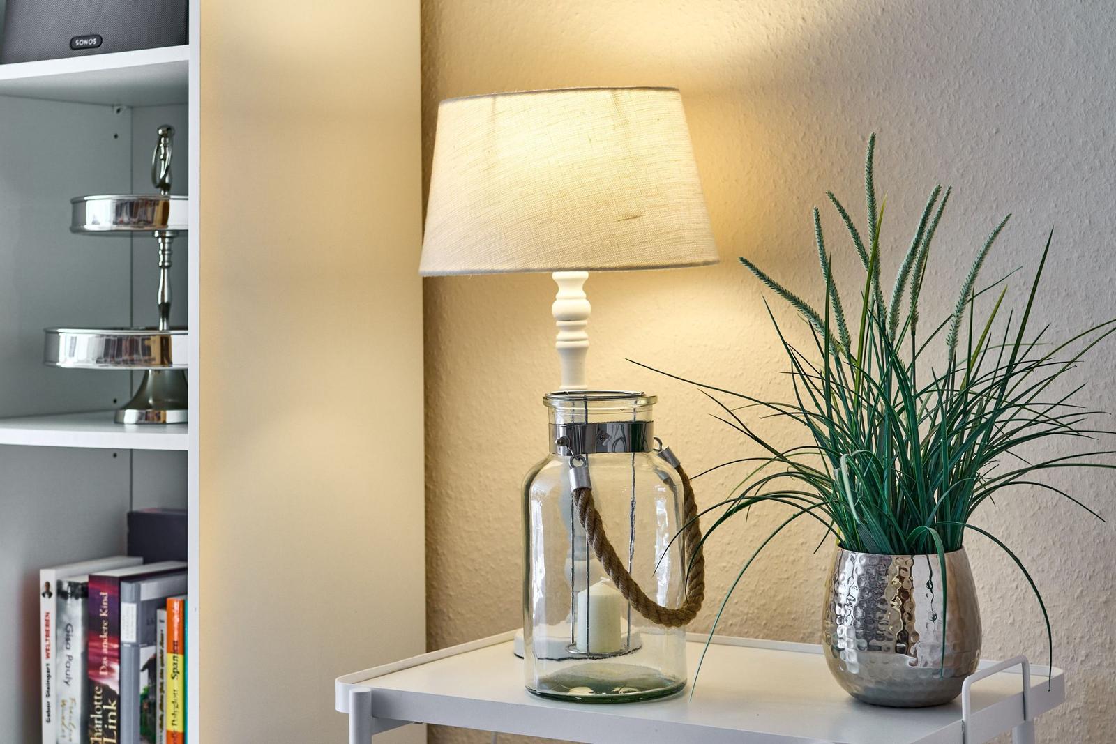 Tischlampe in Glasgefäß mit Seil, Pflanze in Metalltopf und Bücherregal im Hintergrund.