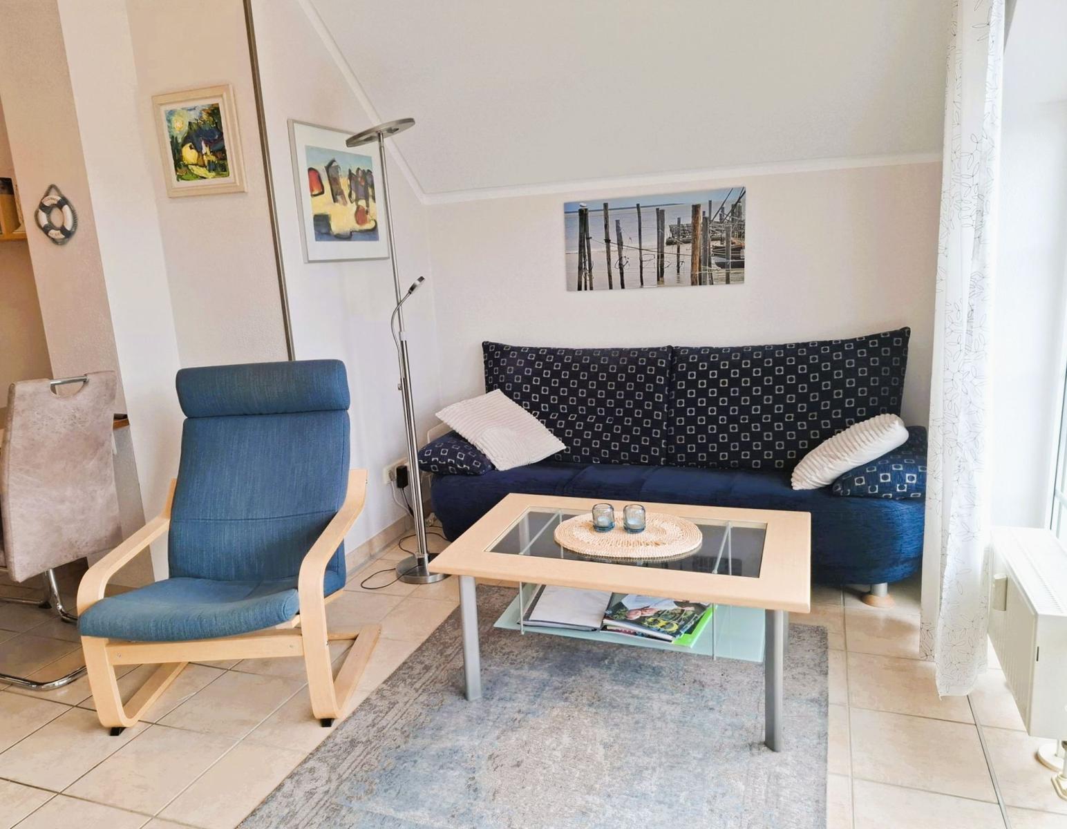 Ein Wohnzimmer mit Sofa, Sessel und Couchtisch. An den Wänden hängen Bilder.