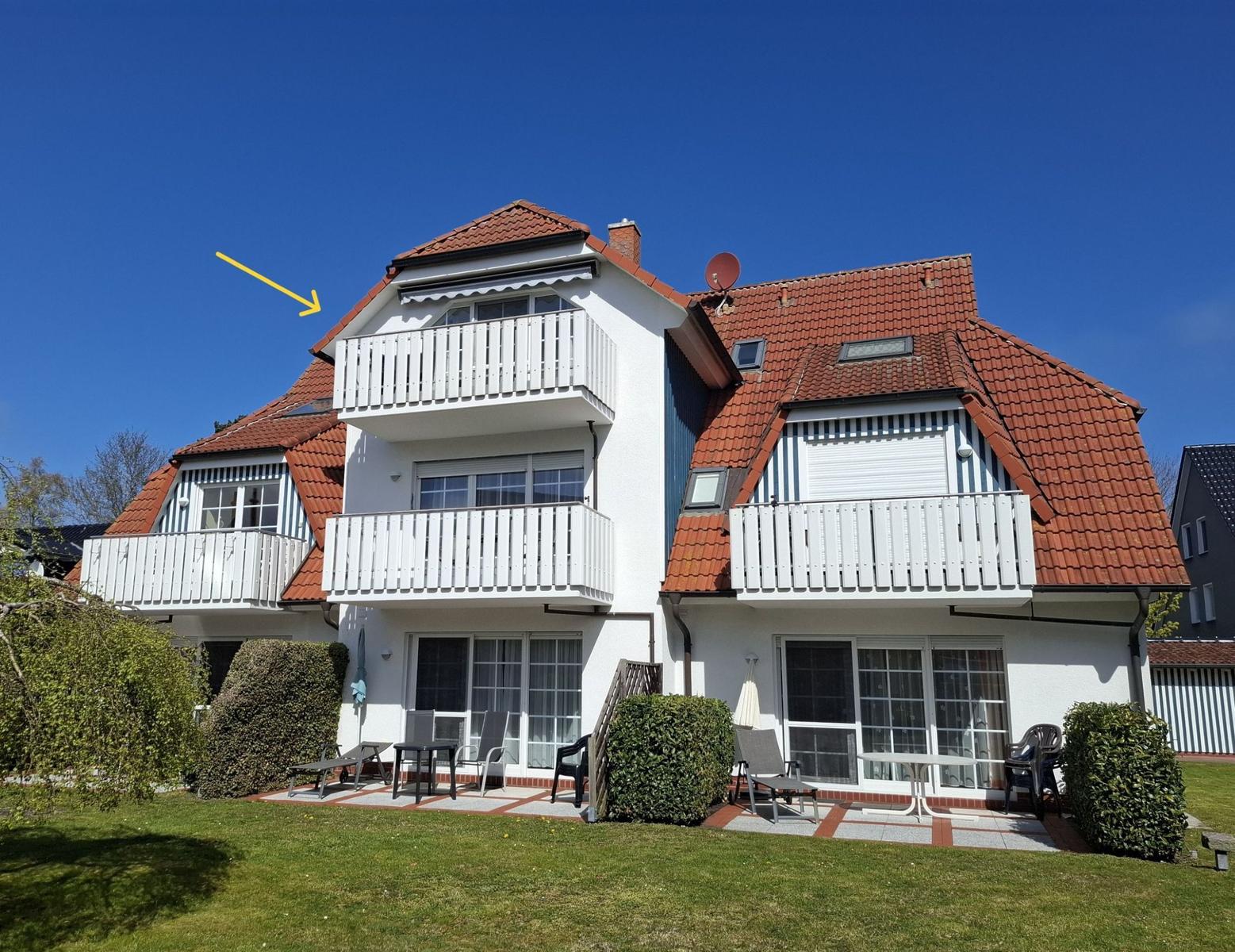 Ferienhaus mit weißen Balkonen, rotem Dach und Gartenbereich mit Sitzgelegenheiten.
