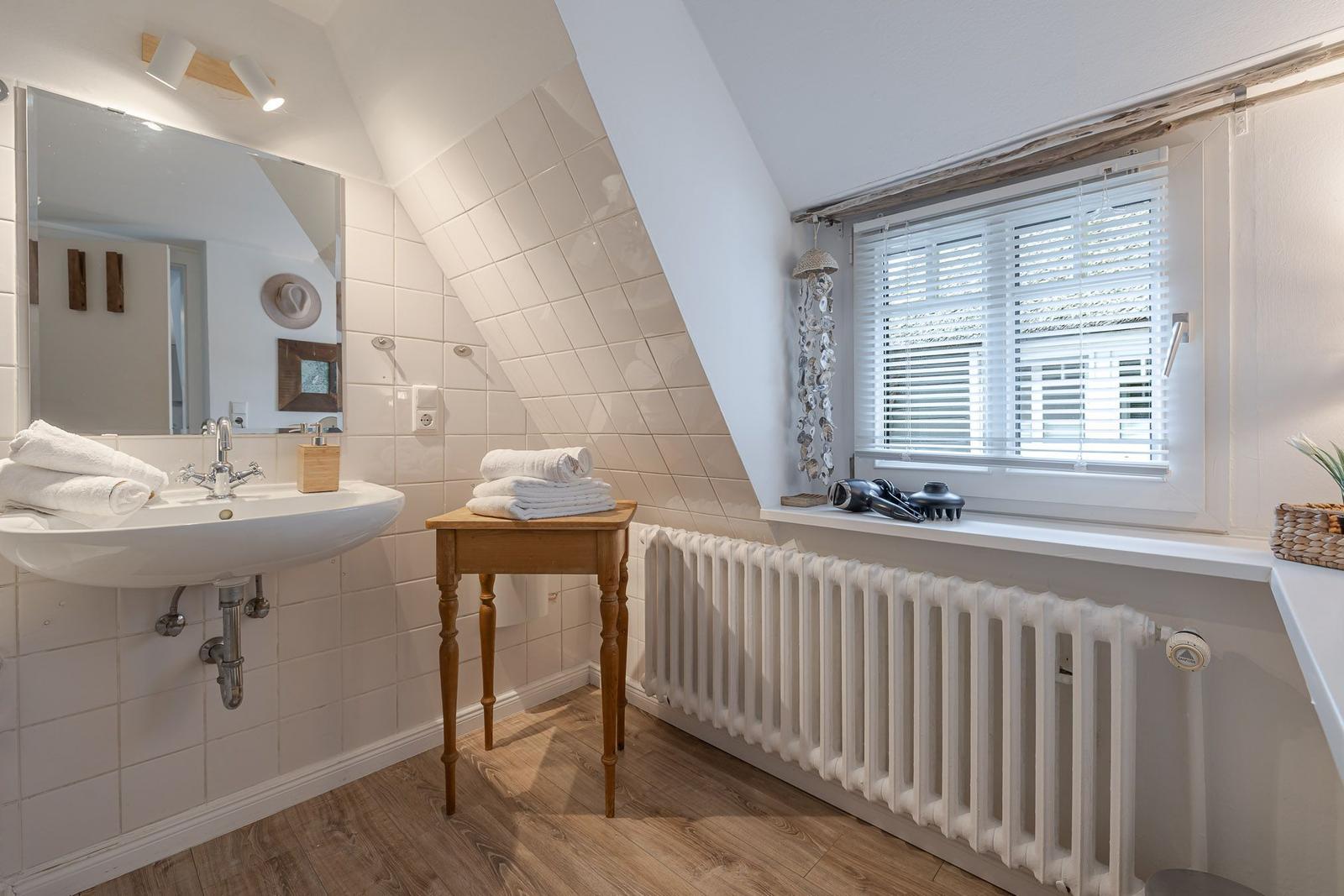 Badezimmer mit Waschbecken, Spiegel, Holztisch und Fenster mit Jalousie.
