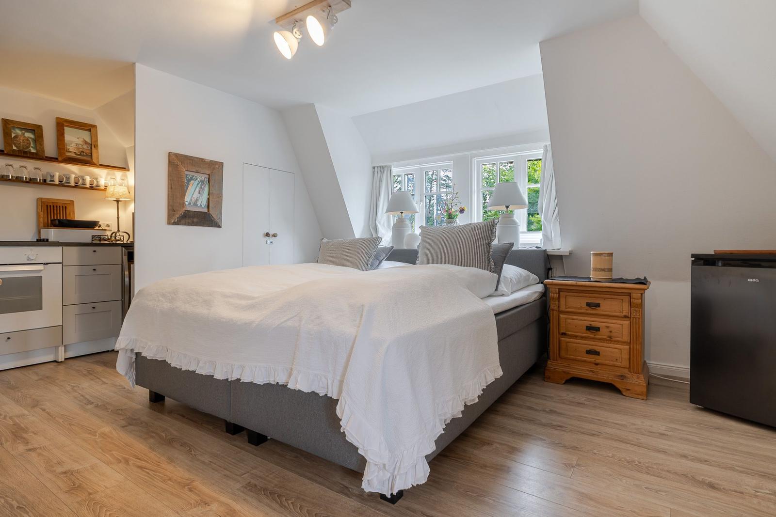 Schlafzimmer mit Bett, Nachttisch und Fenster. Küche und Kühlschrank sind sichtbar.