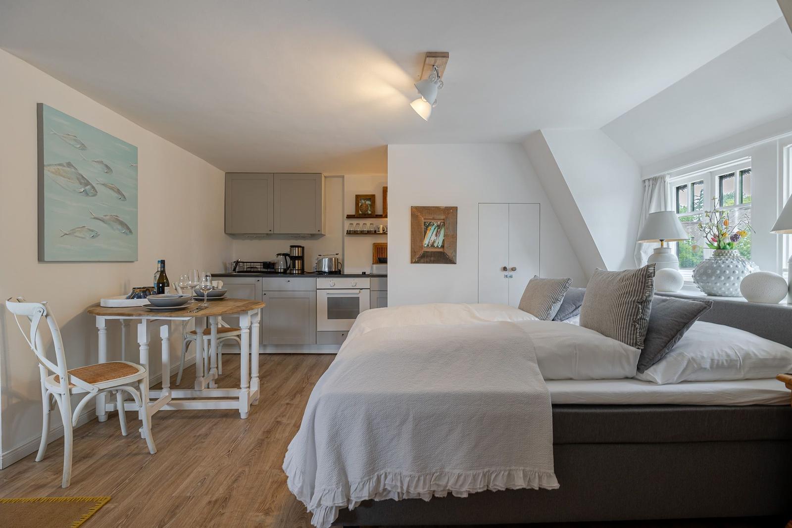 Schlafzimmer mit Bett, Küchenbereich und Essplatz. Fenster mit Blumen und Wandbild.