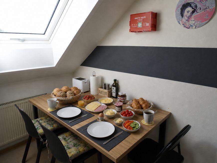 Frühstückstisch mit Brot, Käse, Obst und Getränken unter Dachfenster.
