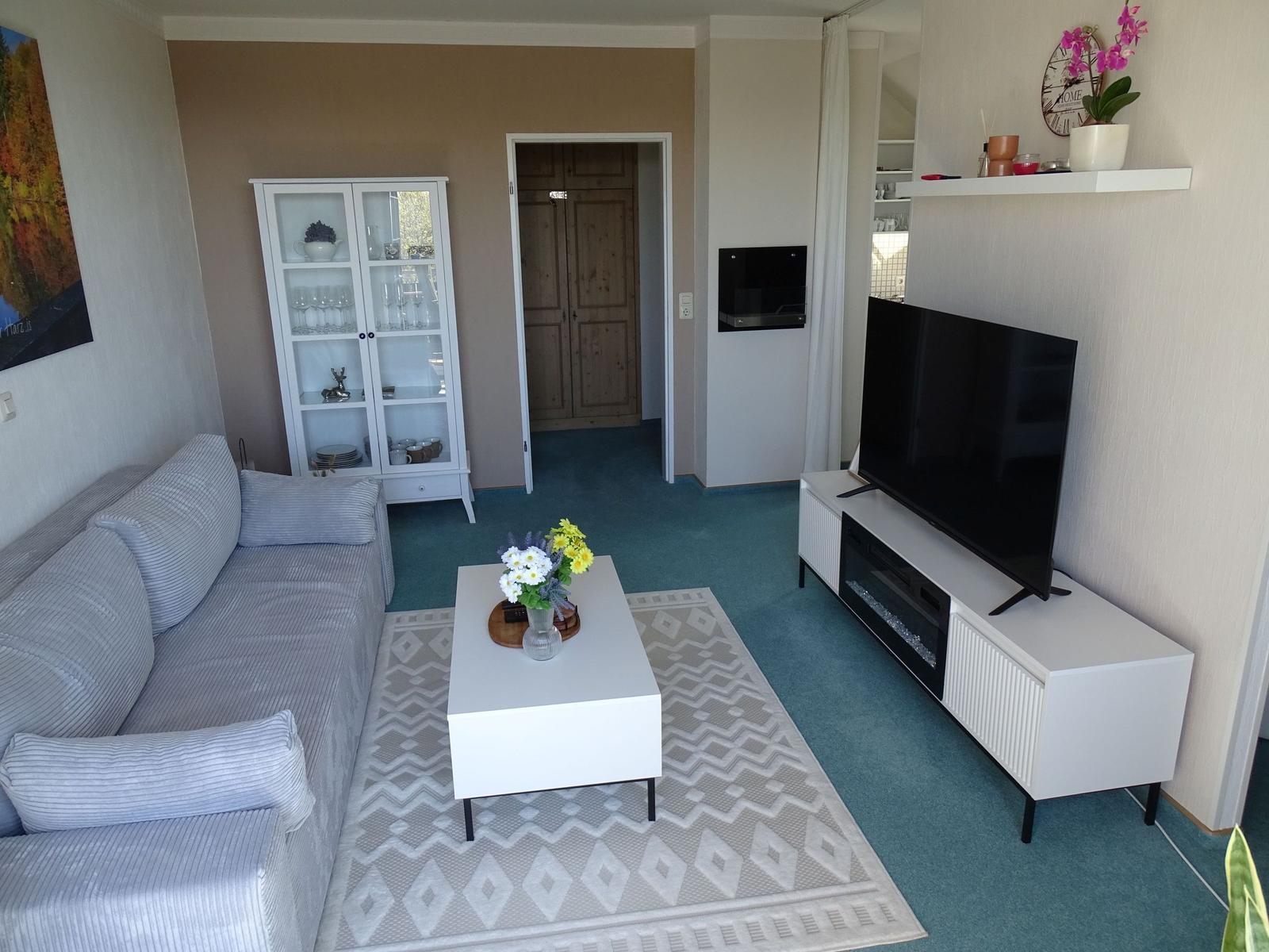 Wohnzimmer mit Sofa, TV und Glasvitrine. Teppich und Couch sind weiß.
