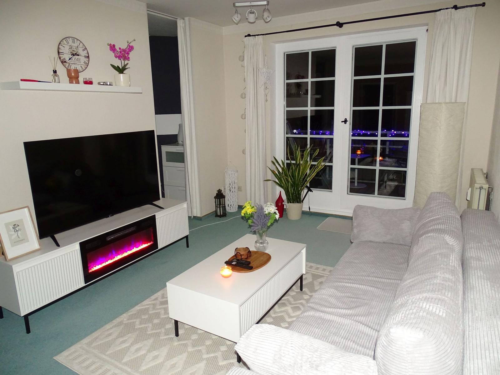 Wohnzimmer mit Sofa, Couchtisch und Fernseher. Durchgang zur Terrasse mit Sicht auf Lichterkette.