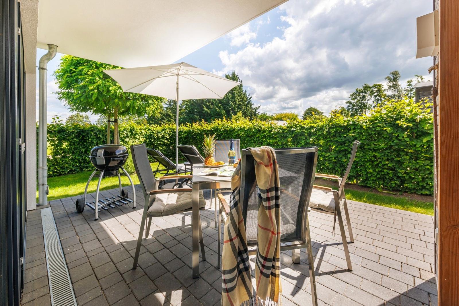 Terrasse mit Tisch, Stühlen, Sonnenschirm und Grill. Grünhedge im Hintergrund.