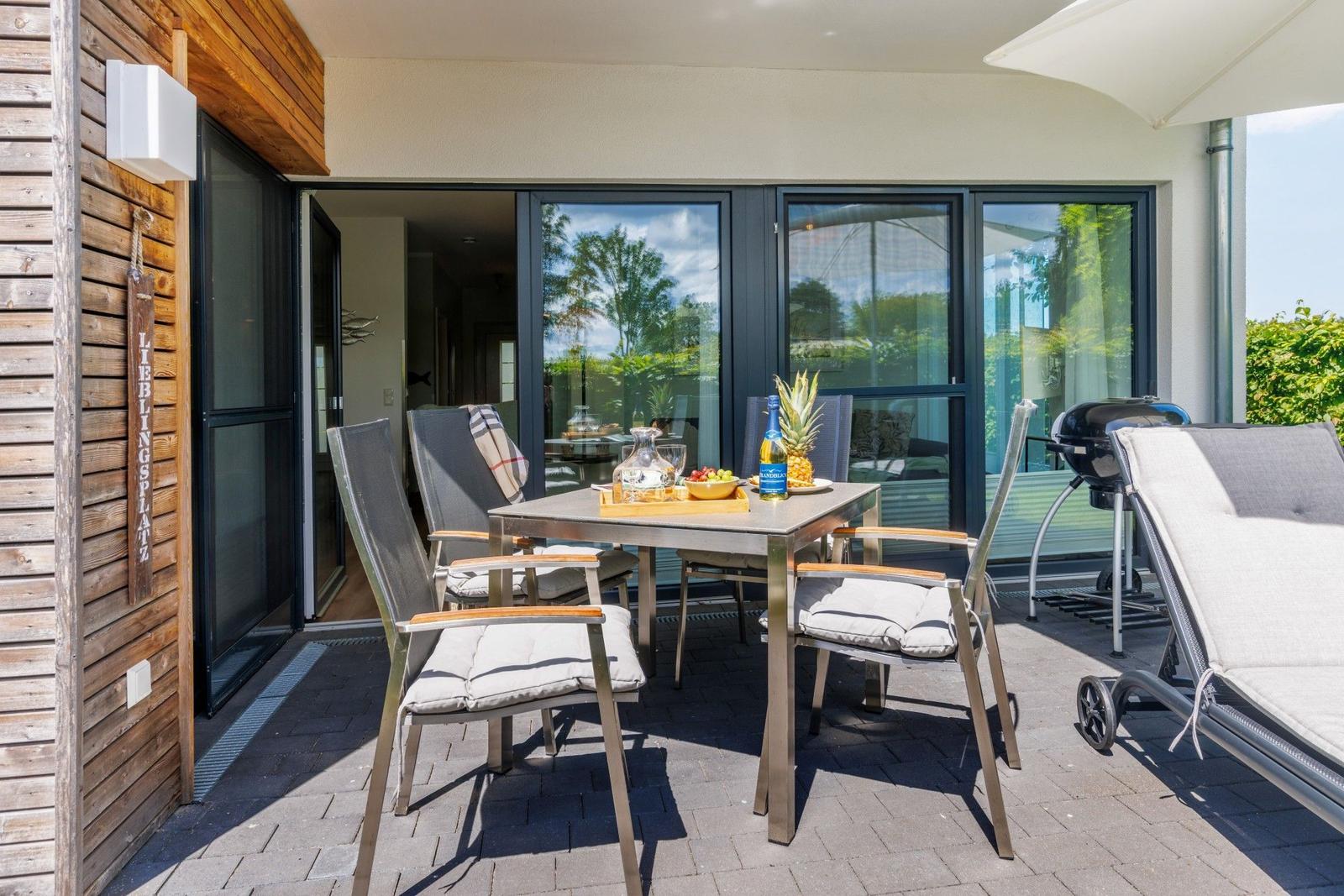 Terrasse mit Tisch, Stühlen und Liege unter Sonnenschutz. Grill und Obst auf Tisch.