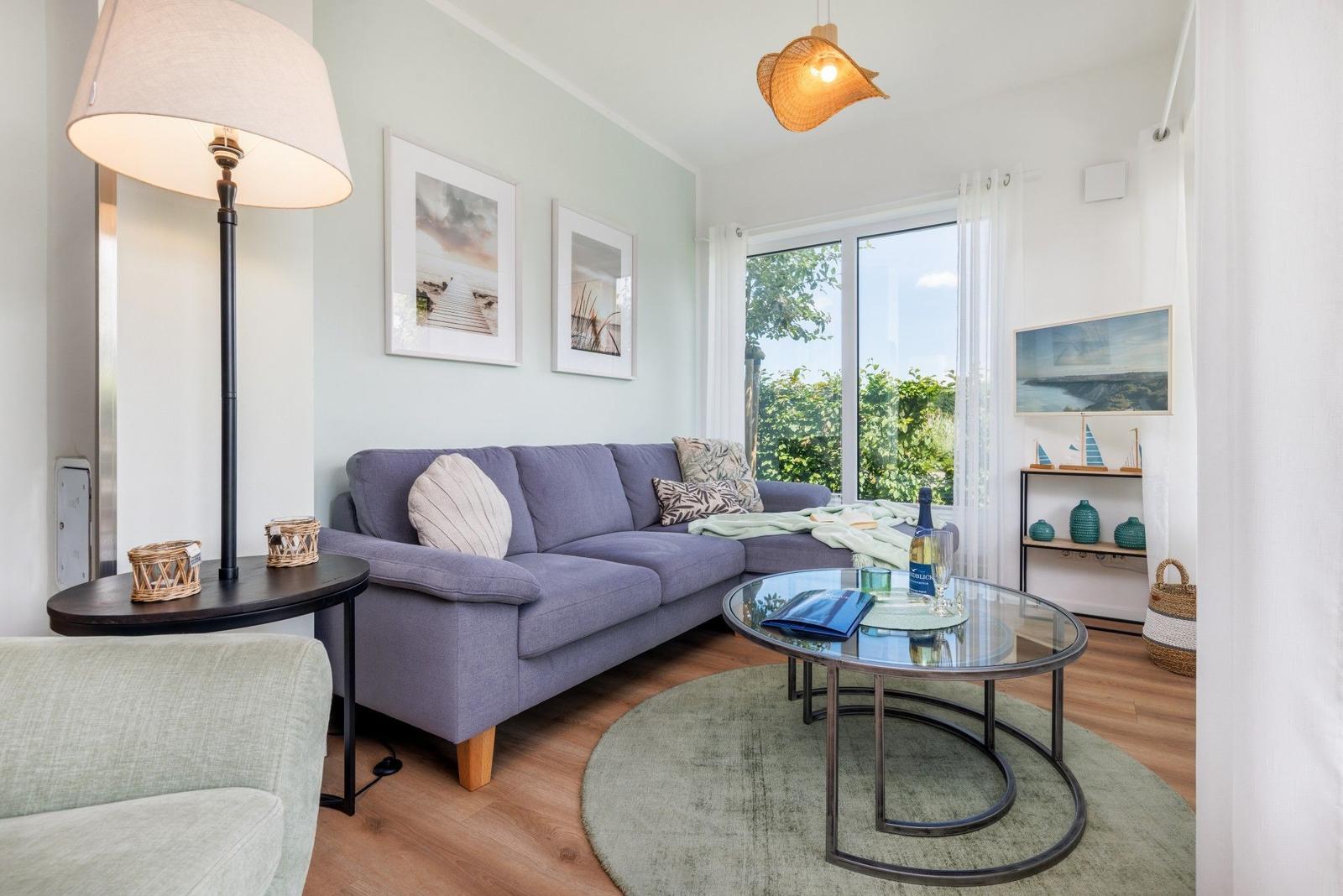 Ein Wohnzimmer mit grauem Sofa, Glas-Tisch und Blick auf Grün.
