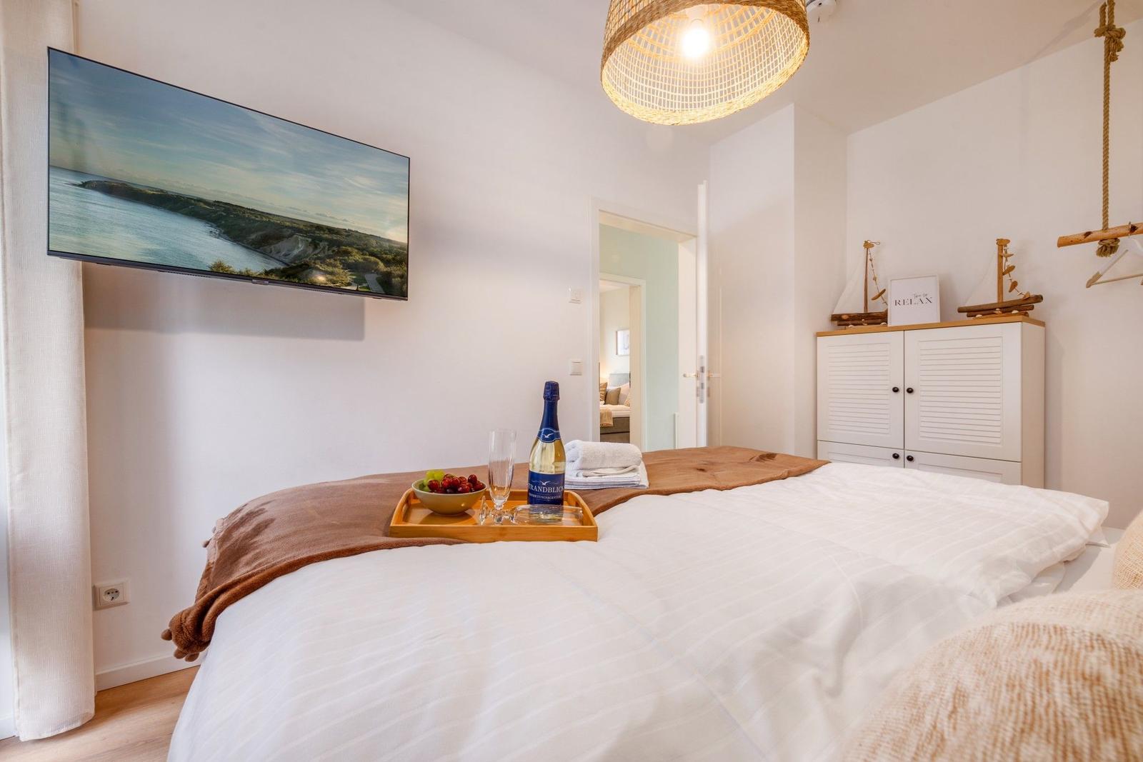 Schlafzimmer mit Bett, Fernseher und Schrank. Auf dem Bett steht ein Tablett mit Wein und Obst.
