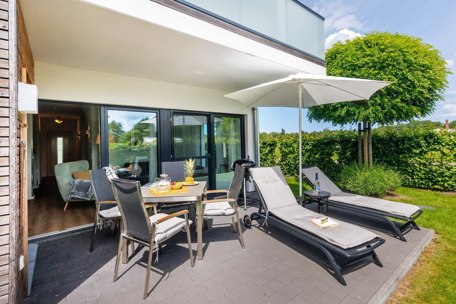 Terrasse mit Liegestühlen, Tisch und Sonnenschirm. Blick in Wohnbereich und grünen Garten.