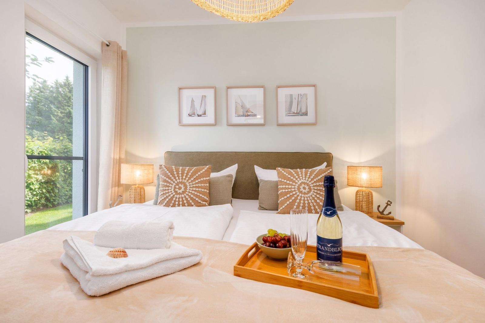Schlafzimmer mit Bett, Fenster und Bildern an der Wand. Auf dem Bett steht ein Tablett mit Wein, Obst und Handtüchern.