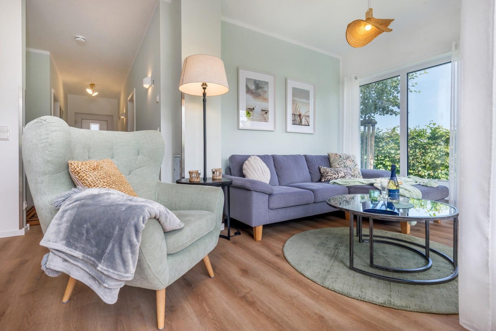 Ein Wohnzimmer mit grauem Sofa, grünem Sessel und Glas-Tisch. Große Fenster mit Blick auf Grünflächen.