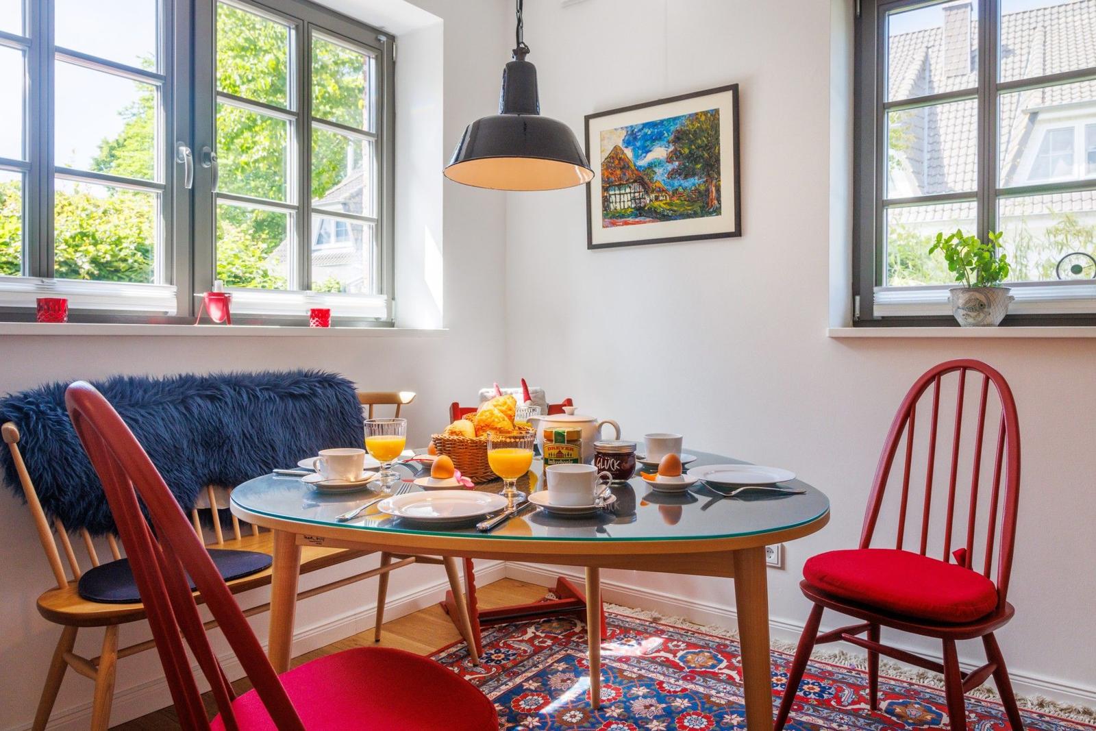Esstisch mit roten Stühlen und Frühstück. Fenster, Teppich und Wandbild.