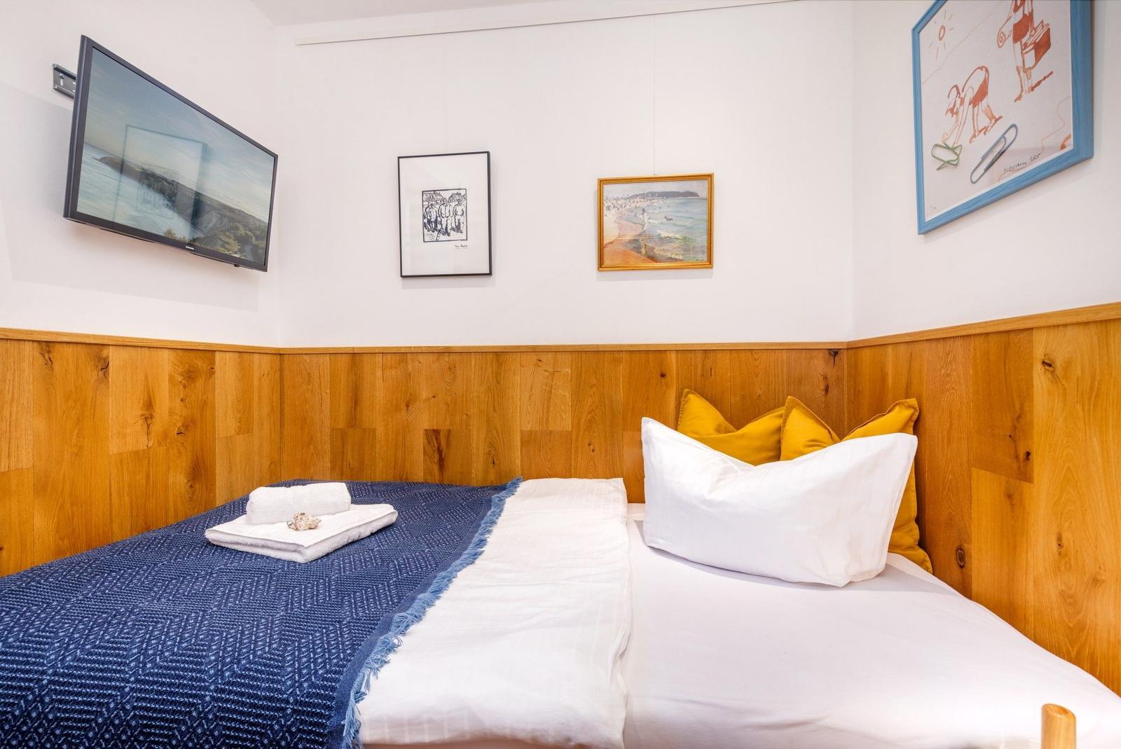 Ein Bett mit weißer Bettwäsche, gelben Kissen und blauer Decke. An der Wand hängen Bilder und ein Fernseher.