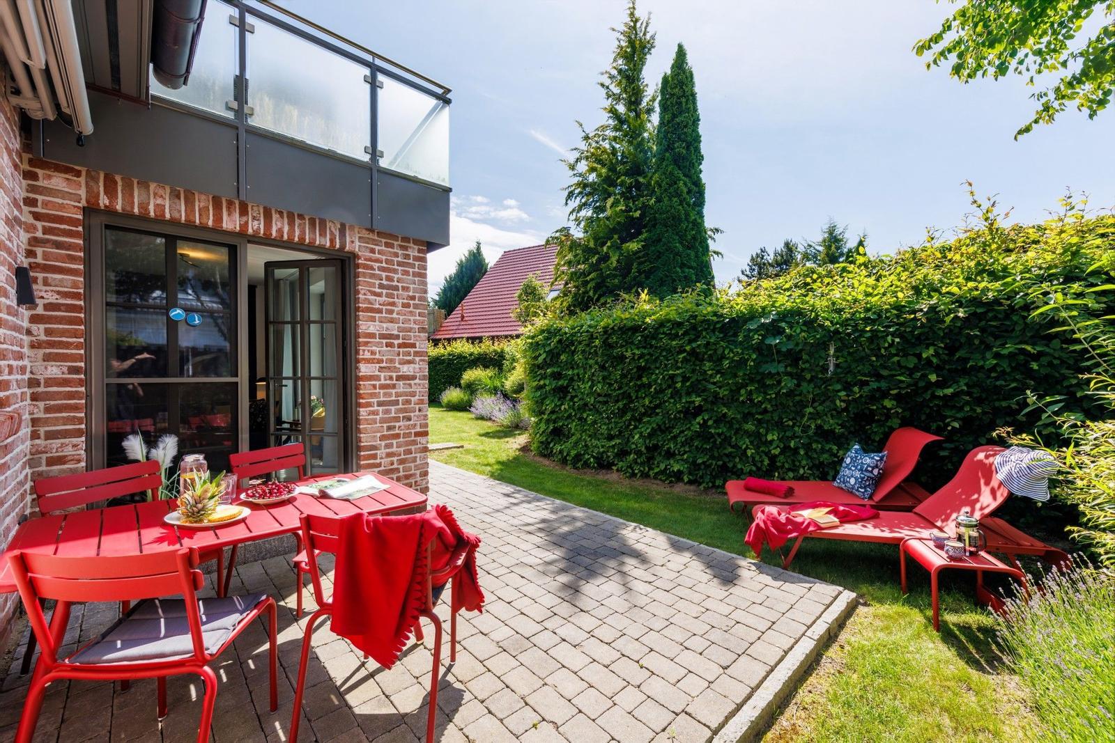 Roter Gartenbereich mit Tisch, Stühlen und Liegen. Hinterhof mit Hecke und Blick auf Haus.