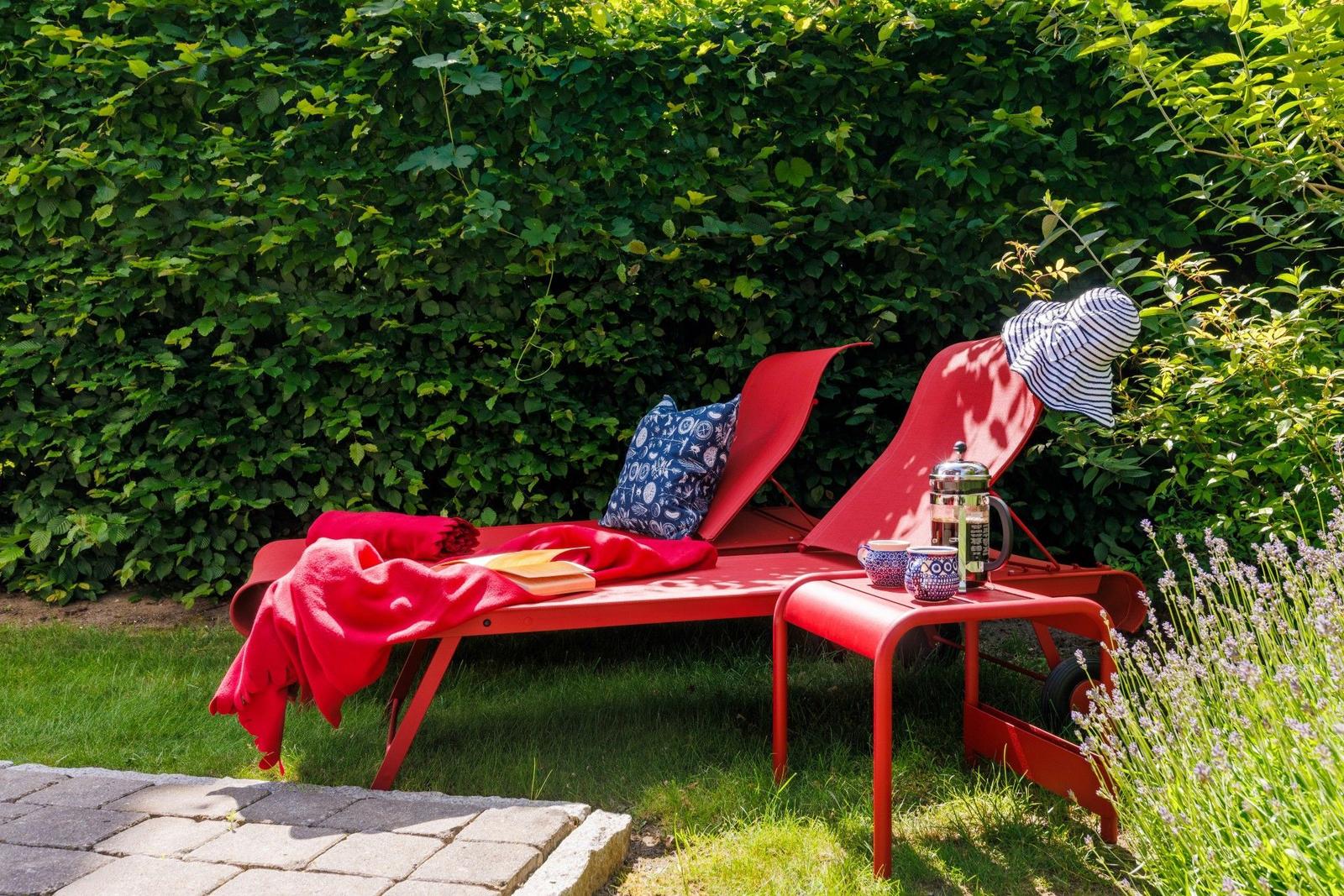 Zwei rote Liegestühle im Garten mit Kissen, Decke und Tisch mit Kanne und Tassen.