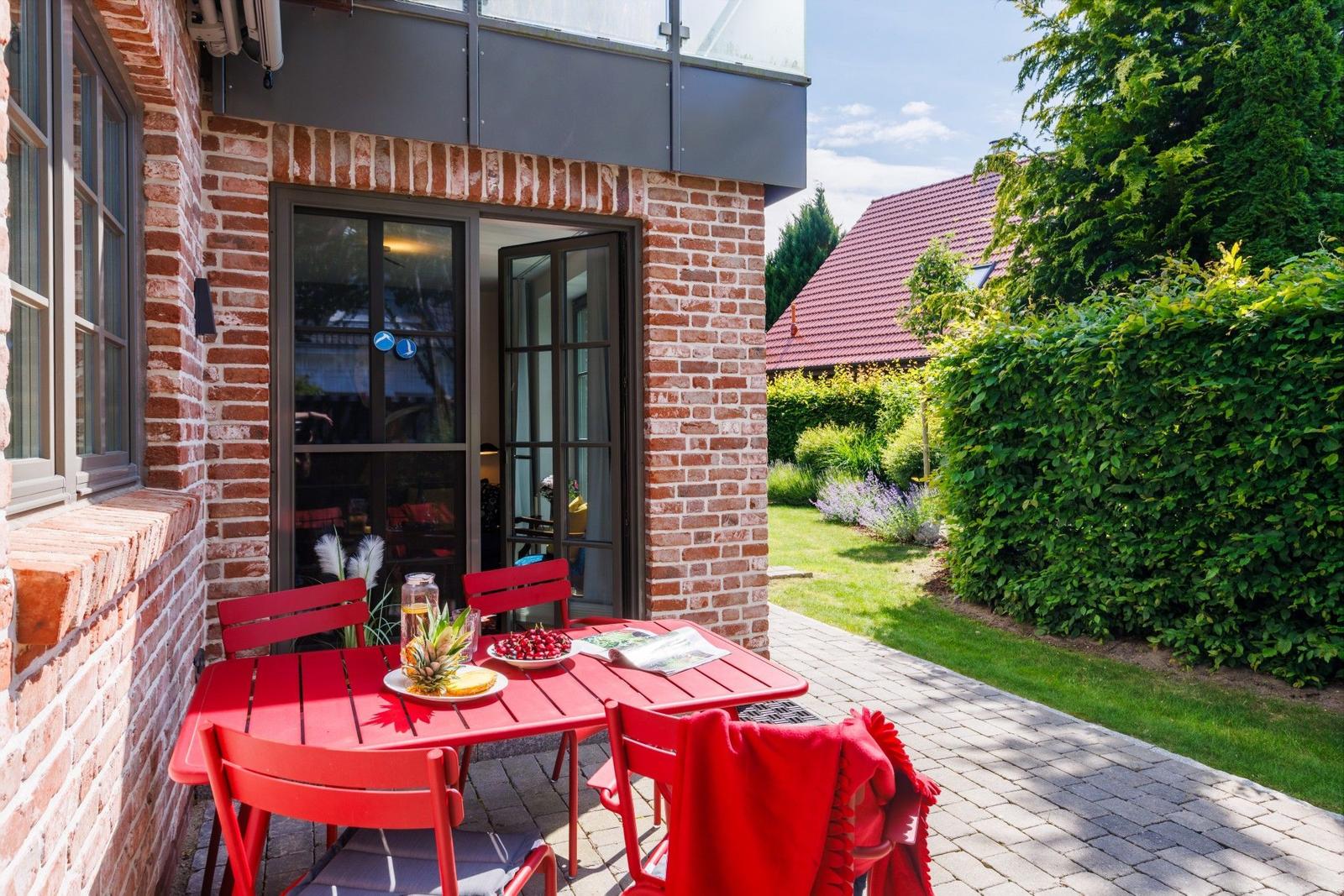 Roter Tisch und Stühle auf gepflastertem Hof vor Backsteinhaus mit Garten.