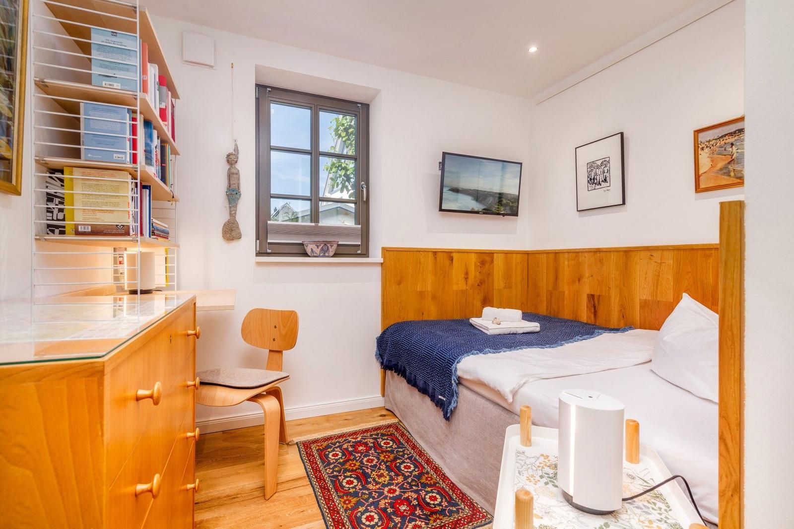 Ein Schlafzimmer mit Bett, Schreibtisch, Bücherregal und Fenster. Wandbild und Teppich sind sichtbar.