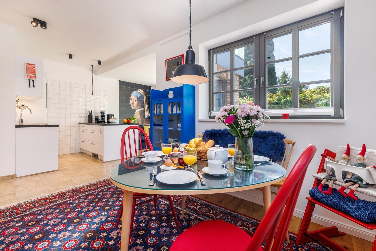 Esszimmer mit Glas-Tisch, roten Stühlen und blauem Schrank. Fenster zeigt Gartenansicht.