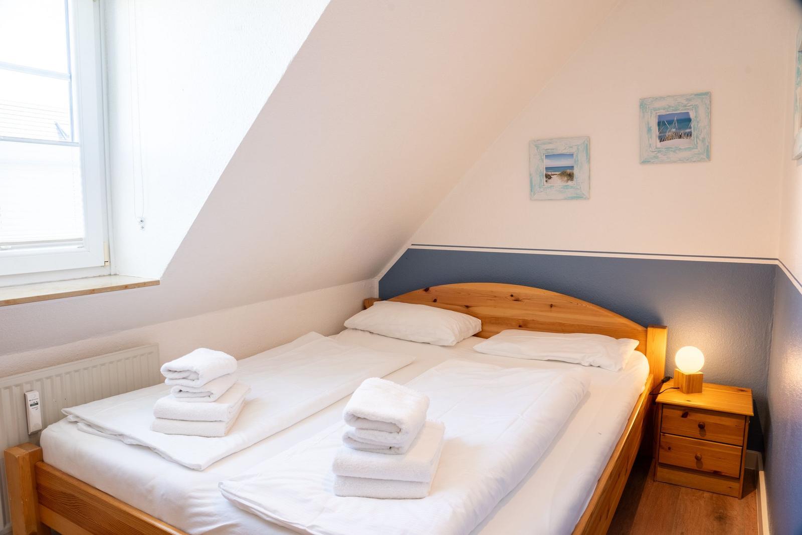 Schlafzimmer mit Dachschräge, Bett mit weißen Laken und Handtüchern, Nachttisch mit Lampe.