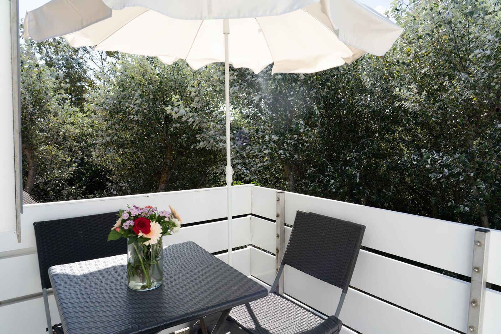 Terrasse mit Tisch, Stühlen und Sonnenschirm. Blumenarrangement und Blick in den Garten.