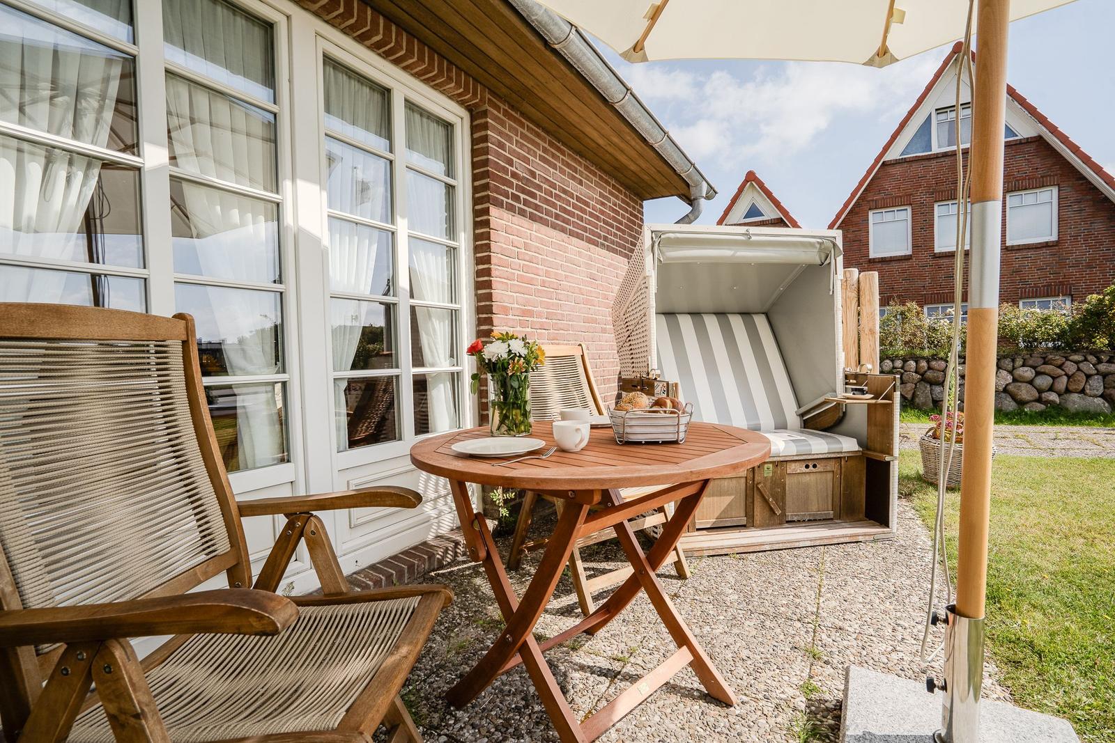 Terrasse mit Holztisch, Stühlen und Strandkorb. Hintergrund: Backsteinhaus und Garten.