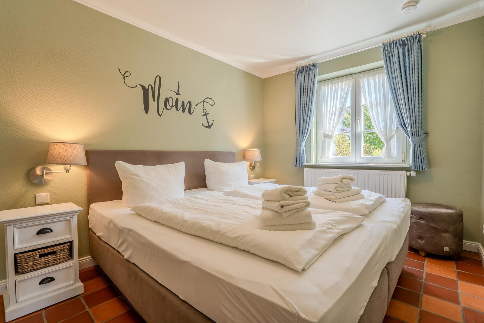 Schlafzimmer mit Bett, Nachttisch und Fenster. Wanddekoration mit Moin und Anker.