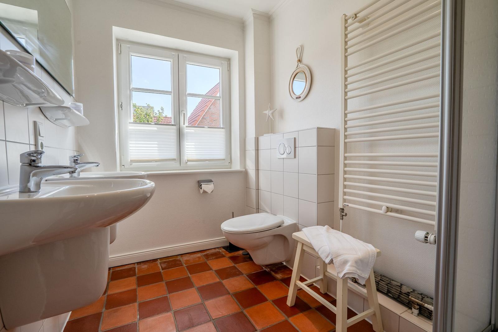 Badezimmer mit Waschbecken, Toilette und Fenster. Terrakottafußboden und Heizkörper.