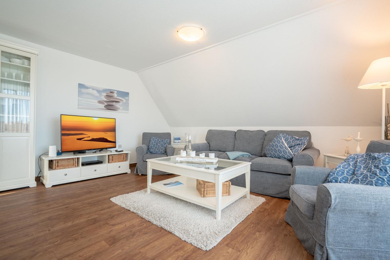 Wohnzimmer mit grauem Sofa, weißem Couchtisch und Fernseher auf weißen Möbeln.
