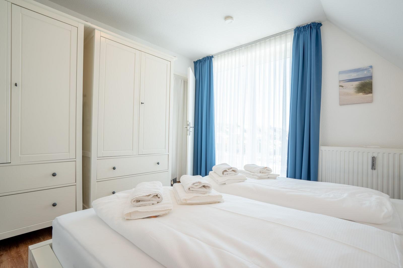 Zwei Betten mit weißen Laken, weiße Schränke, blaue Vorhänge und ein Fenster.