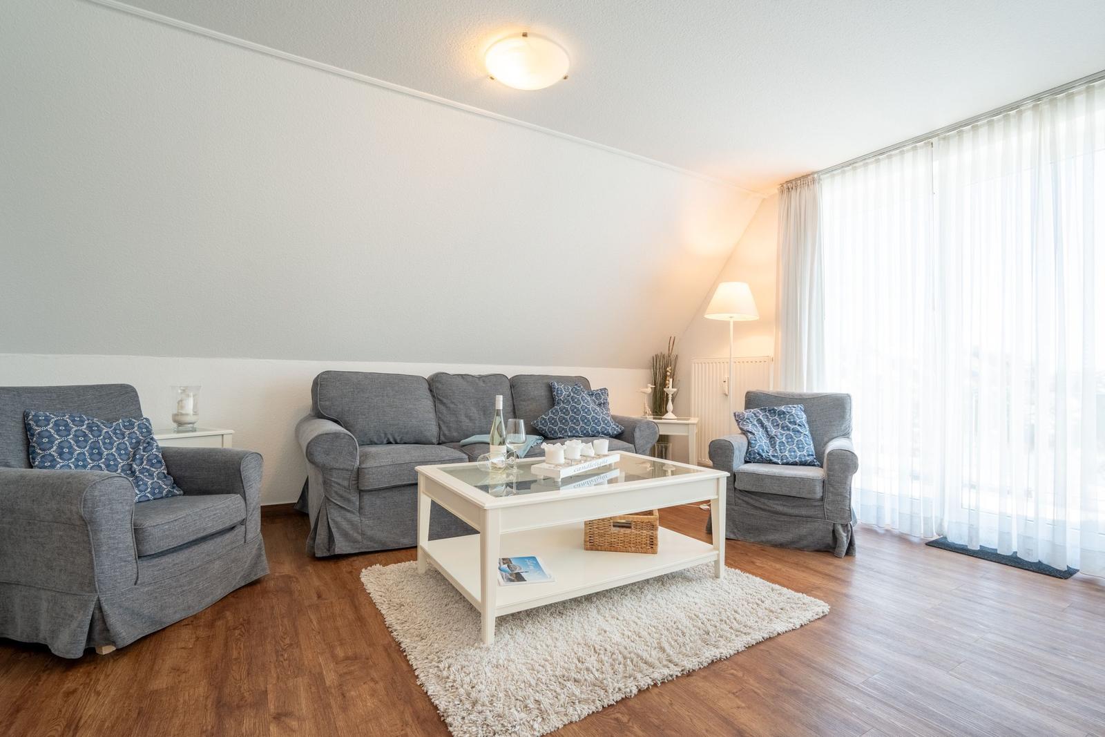 Wohnzimmer mit grauer Couch, zwei Sesseln, weißem Tisch und Holzboden.