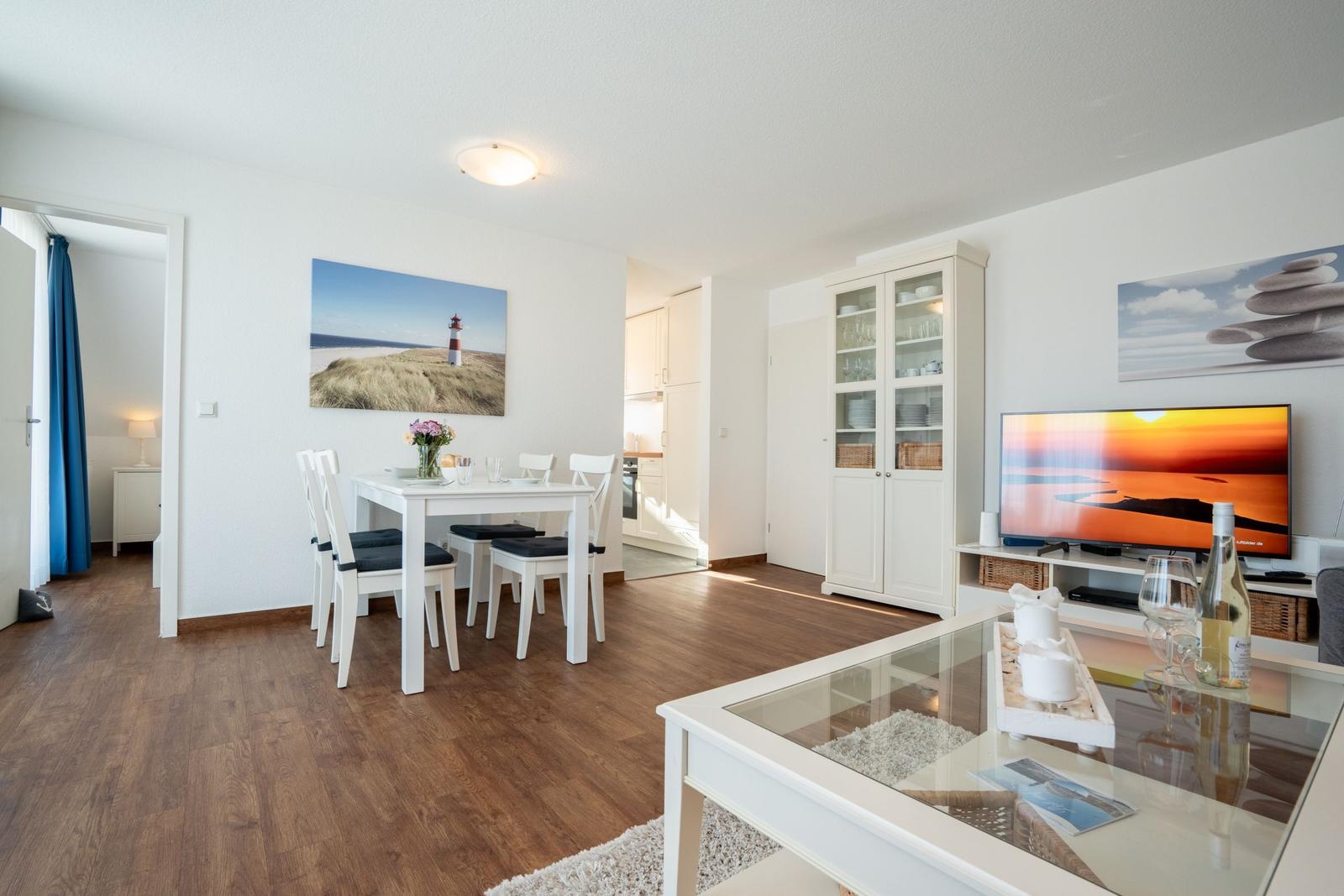 Wohn- und Essbereich mit weißem Tisch, Glaswand-Tisch und TV. Küche und Schlafzimmer sind sichtbar.