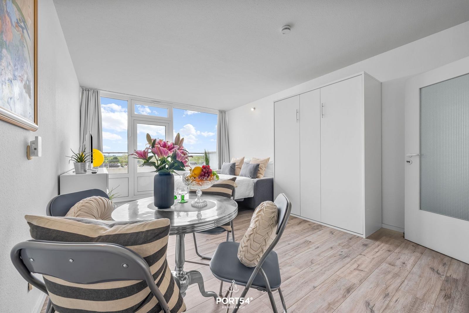 Dining area with table, chairs, and view to balcony. Background: sleeping area with bed and wardrobe.