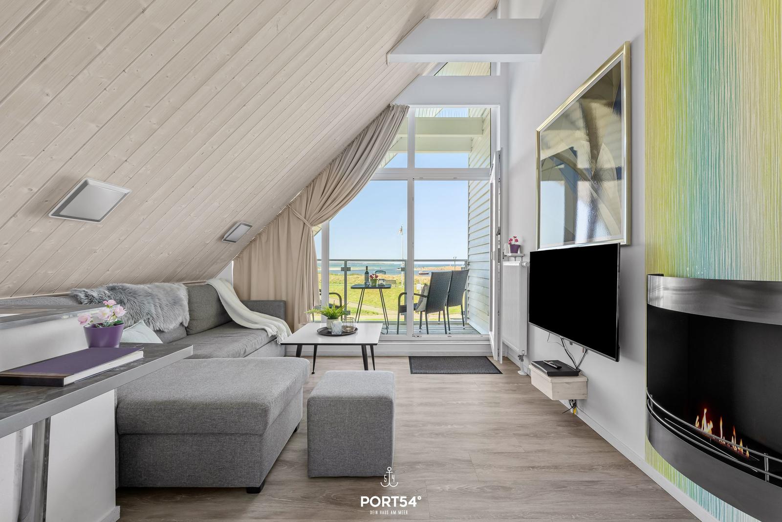 Wohnbereich mit Sofa, Fernseher und Kamin. Balkon mit Meerblick. Holzdecke und Lichtdurchflutung.