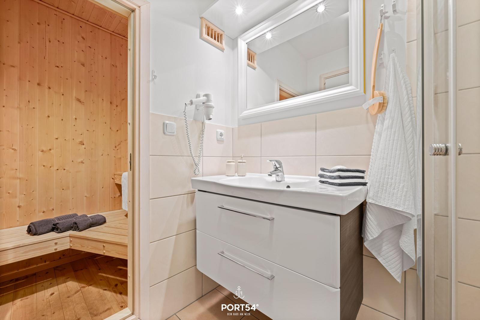 Badezimmer mit Waschbecken, Spiegel und Dusche. Sauna mit Holzwand und Bänken.