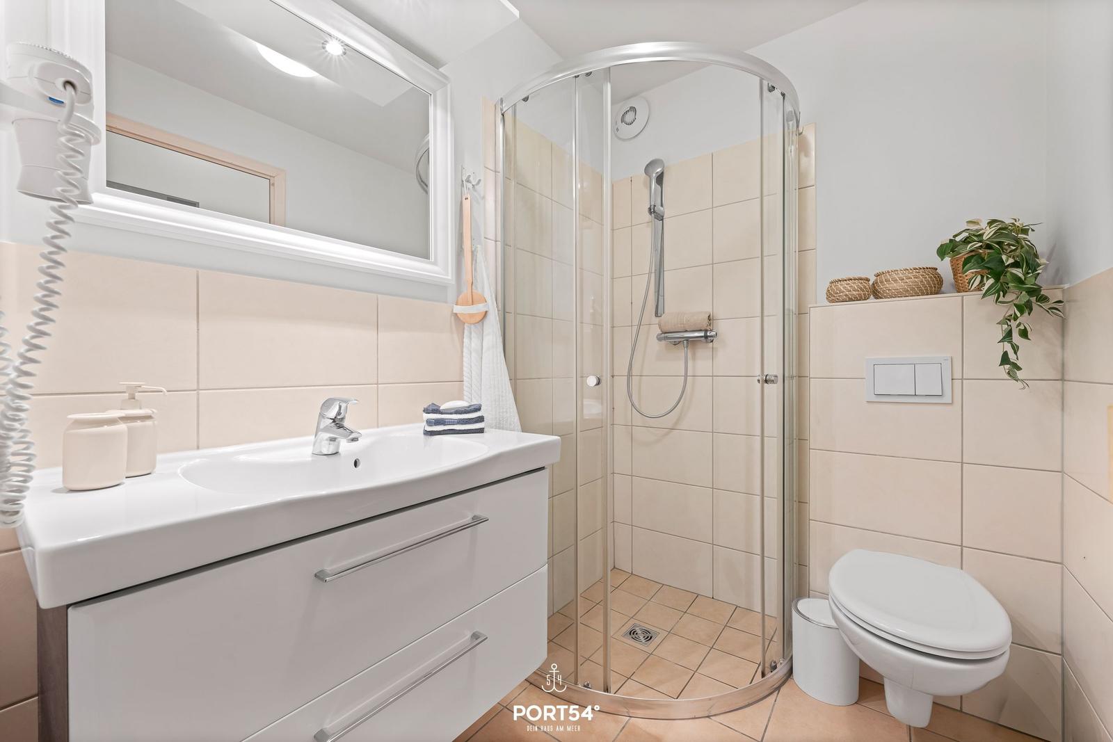 Badezimmer mit Dusche, Waschbecken und Toilette. Weiße Einrichtung und beige Fliesen.